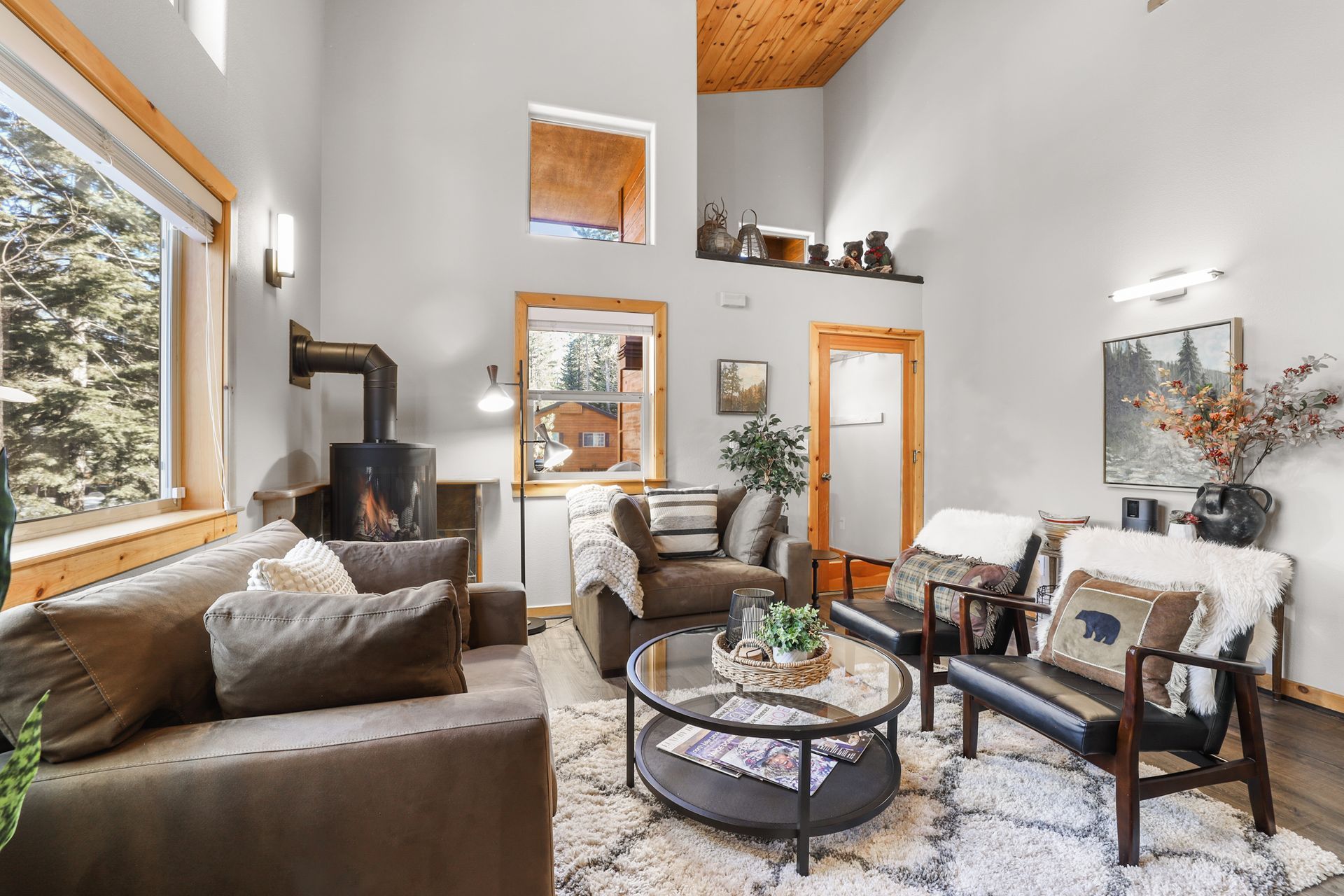 Cozy living room with sofa, chairs, coffee table, wood stove, and large window overlooking trees