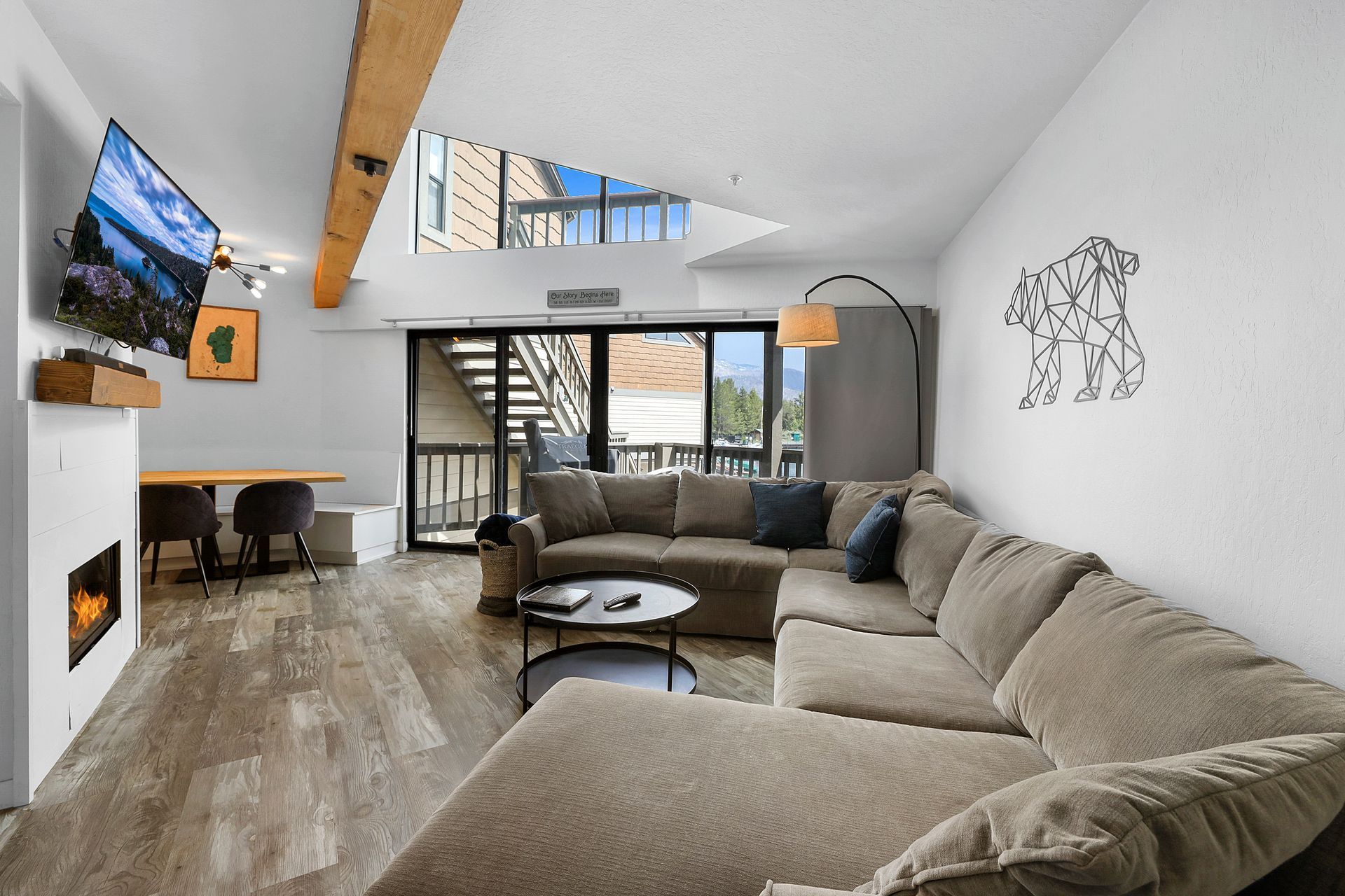 Modern living room with beige sectional, wall-mounted TV, fireplace, and staircase under a skylight
