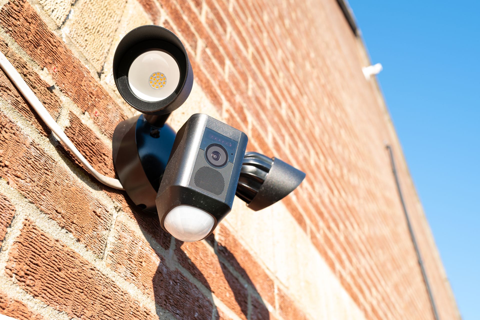 Security light mounted on a brick wall, angled upward in bright sunlight.