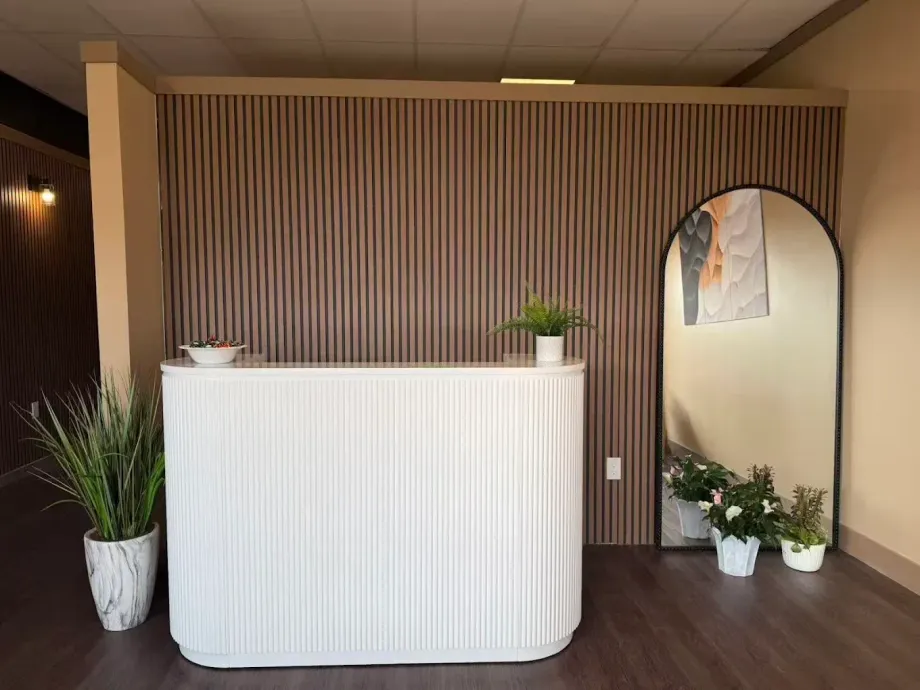 Reception desk with white front, wood slat wall, arched mirror, and potted plants in a warm lobby