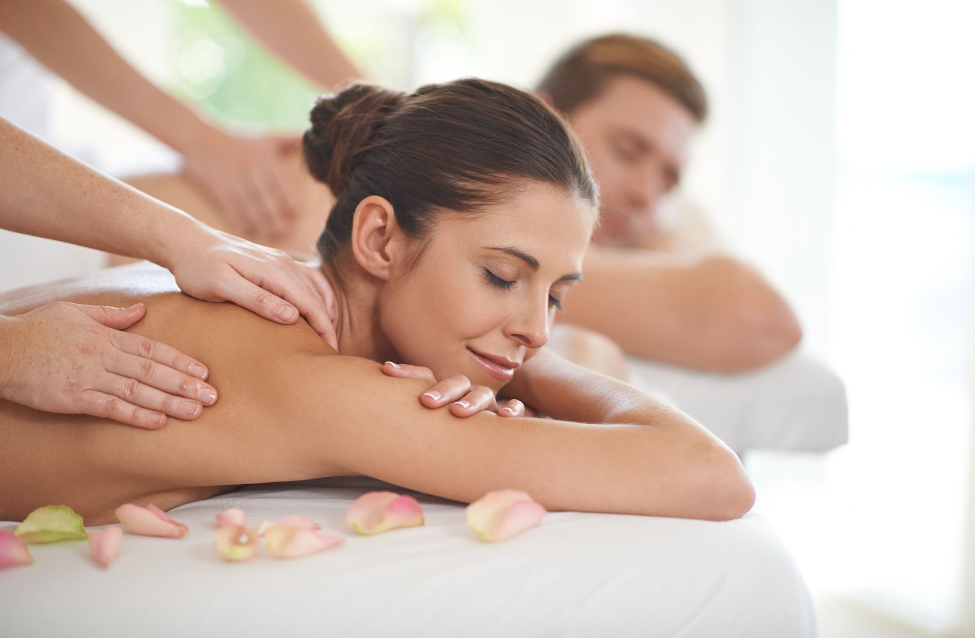 Spa massage with woman resting face down while two therapists massage her shoulders and back with flowers nearby