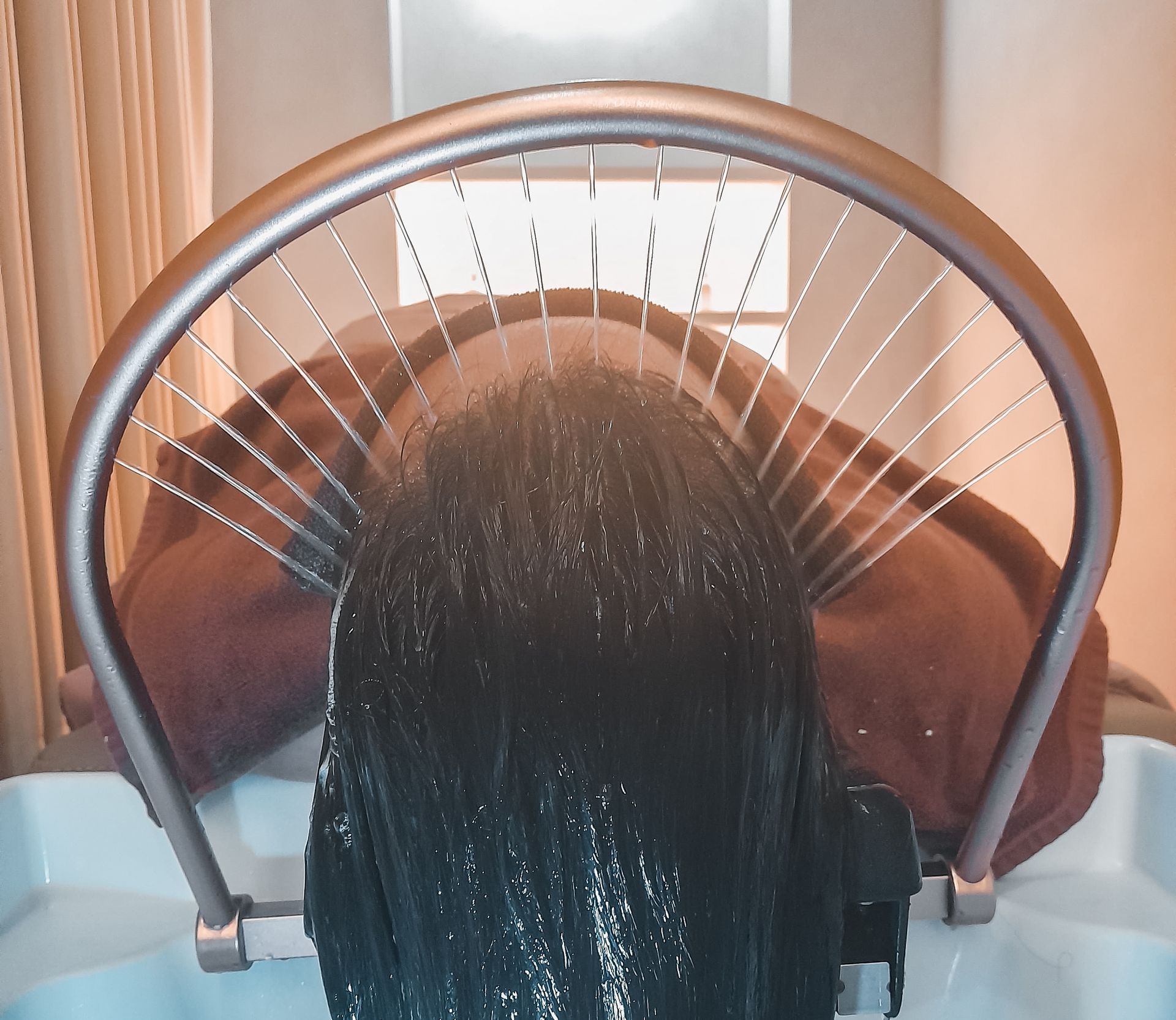 Person seated under a hair-washing sink with water spraying onto long dark hair