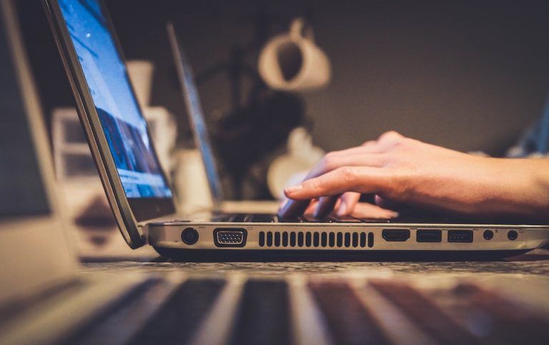 Hands typing on a laptop keyboard in a dimly lit room, side view of the screen and ports visible