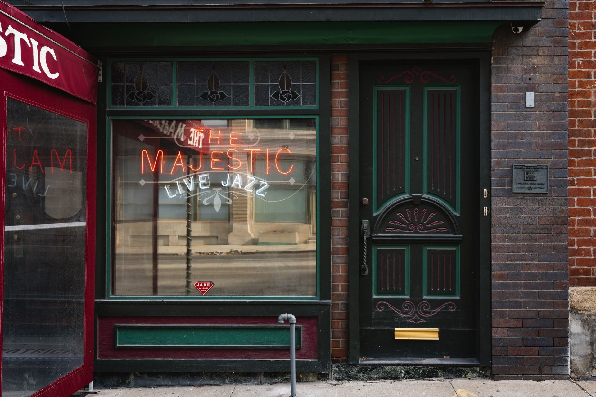 Dark storefront with green trim, large window, and door, next to a red awning on a brick street.