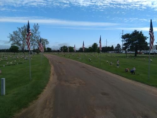 Dirt road curving through a cemetery with American flags and headstones under a blue sky