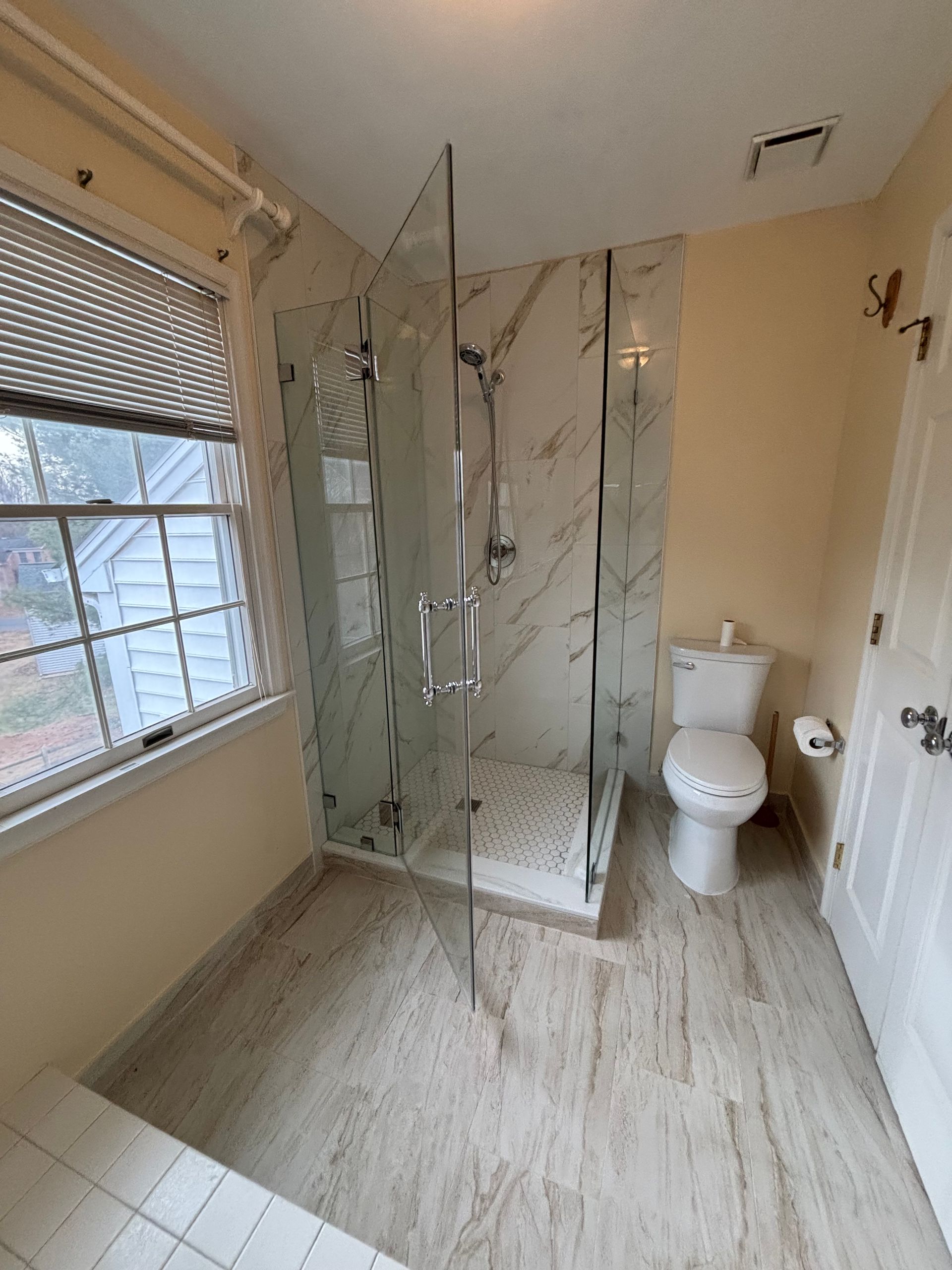 Bathroom with marble tile, glass shower, toilet, window with blinds, and white door.