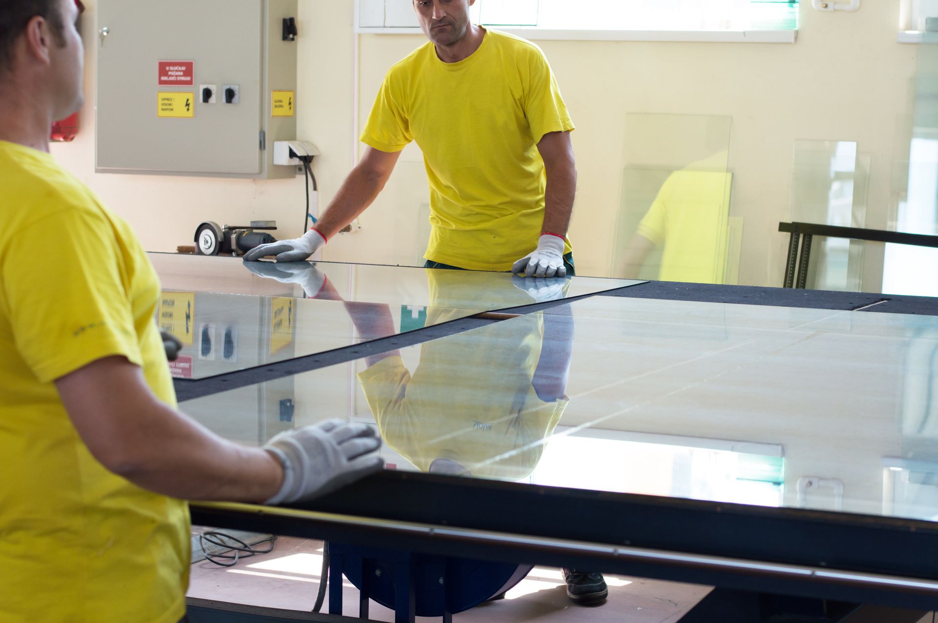 Two men in yellow shirts are working on a large piece of glass.