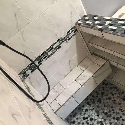 A walk-in shower with white marble-look tiles, a mosaic accent strip, built-in shelving, a bench, and a pebble floor.