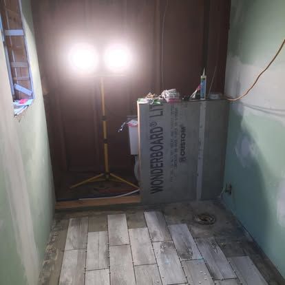 A room under renovation with light-colored tile flooring, a work light, and a piece of WonderBoard leaning against a wall.