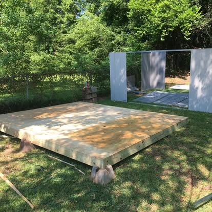 A wooden shed base sits on concrete blocks in a grassy yard, with unfinished shed wall panels standing in the background.