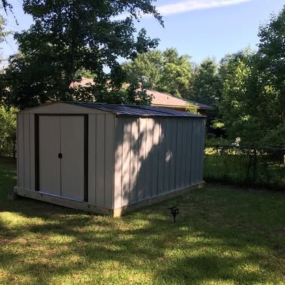 A light grey metal storage shed with double doors sits on a wooden foundation in a grassy backyard near some trees.