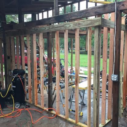 A wooden wall frame under construction with vertical studs, horizontal beams, and an electrical outlet in an open shed.