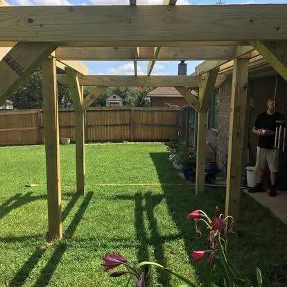 Wooden pergola frame under construction in a grassy backyard next to a house, with a person standing near the structure.