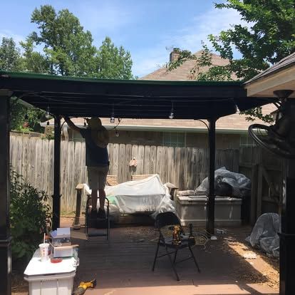 A person stands on a ladder under a dark metal pergola, installing outdoor string lights in a backyard.