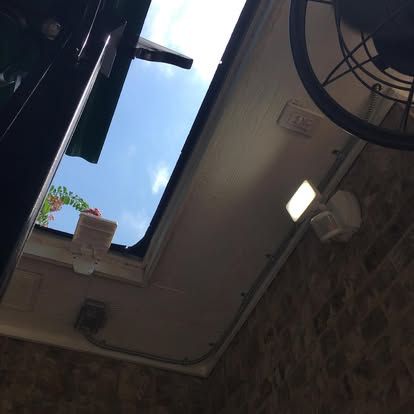 A low-angle view of a partially open skylight showing blue sky, with a motion-sensor security light and a fan mounted nearby.