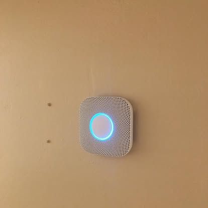 A white Nest Protect smoke and carbon monoxide detector mounted on a beige wall, glowing with a pulsing blue light.