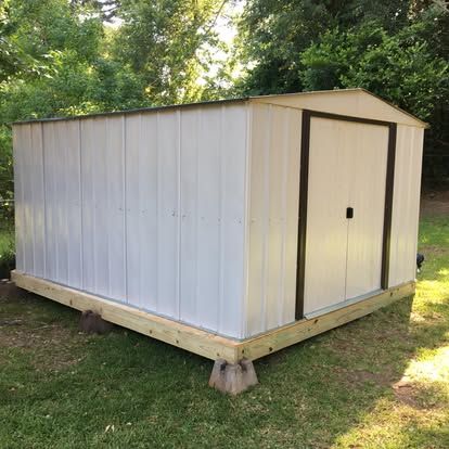 A white metal storage shed with dark brown trim, mounted on a wooden base supported by concrete blocks in a grassy yard.