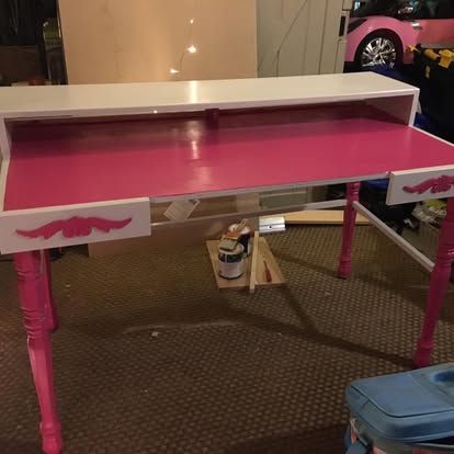 A desk with a bright pink surface, white top shelf, and pink decorative wooden legs in an indoor setting.