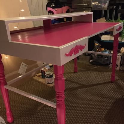 A white and hot pink desk with turned legs and decorative floral appliques on the drawer fronts, set on carpet.
