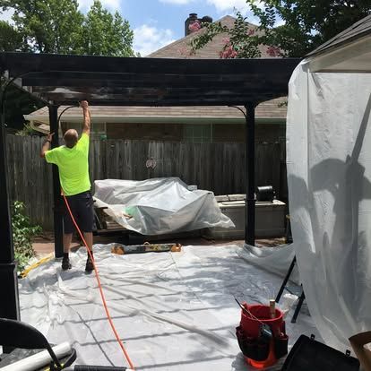 A person in a bright neon shirt uses a spray painter on a black pergola structure covered by plastic sheeting outdoors.