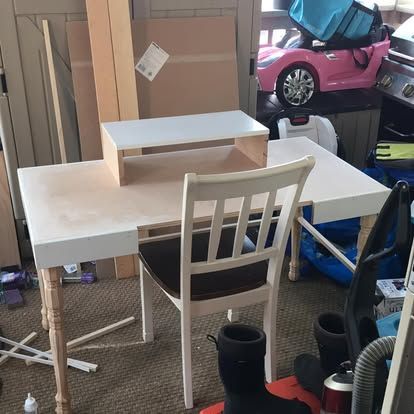 A white desk with a wooden shelf sits in a cluttered room next to a white wooden chair and a pink toy car.