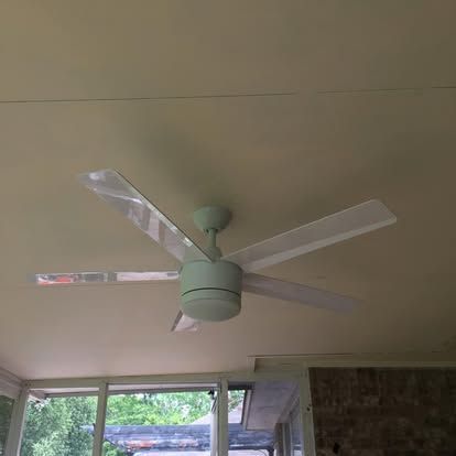 A light-colored four-blade ceiling fan mounted on a plain ceiling above a windowed wall and brick surface.
