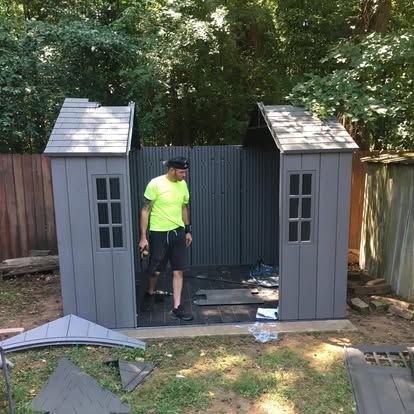 A person in a neon yellow shirt stands inside a partially assembled gray storage shed in a backyard.