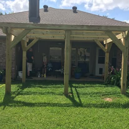 A wooden pergola structure built against the back of a brick house with a grassy lawn in the foreground.