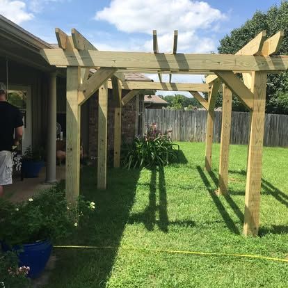 An unfinished wooden pergola stands in a grassy backyard next to a house under a partly cloudy sky.