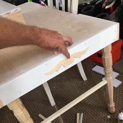 A person aligns a light wood decorative scrollwork appliqué onto the painted white apron of a table frame.