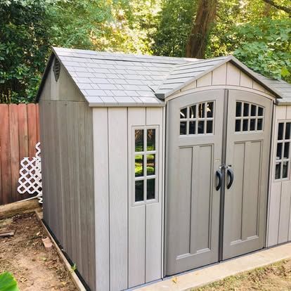 A grey plastic storage shed with double doors, windows, and a shingled roof, situated in a backyard near a wooden fence.