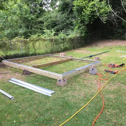 A wooden shed foundation frame sits on concrete blocks in a grassy yard, with construction tools nearby.