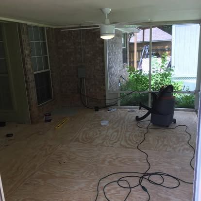 A screened-in porch with plywood flooring, a ceiling fan, a brick wall, and a shop vacuum on the floor.