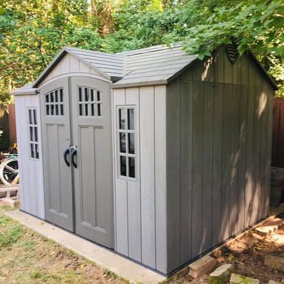 A grey, multi-windowed plastic storage shed sits on a concrete foundation in a wooded backyard.