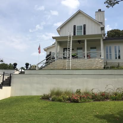 A white, two-story farmhouse with a porch, stone base, tall ladder against the front, and an American flag on the lawn.