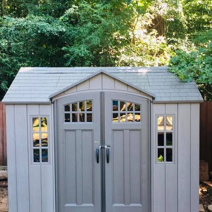 A light gray, resin garden shed with a shingled roof, double doors, and rectangular windows, situated in a wooded area.