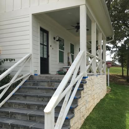 A white farmhouse porch with dark stone stairs, white railings, and a black front door overlooking a grassy lawn.