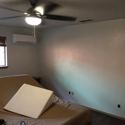 A bedroom with a tan bed, a white foam wedge pillow, a ceiling fan, and a wall-mounted AC unit.