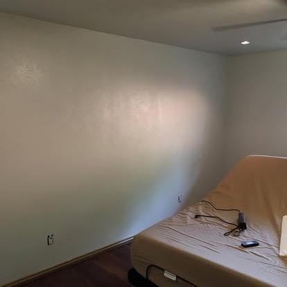A tan-covered hospital-style adjustable bed sits in the corner of a sparsely furnished room with plain, light-colored walls.