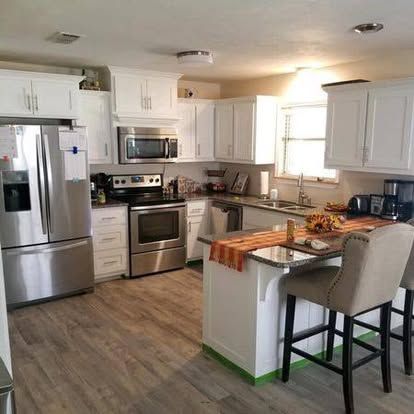 A modern, well-lit kitchen with white cabinets, stainless steel appliances, wood-look flooring, and a breakfast bar.