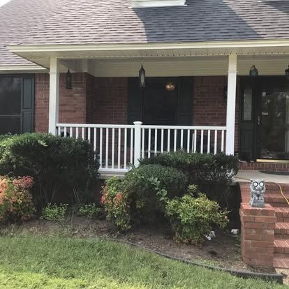 A brick house with a covered front porch, white railing, dark shutters, and landscaping in the front yard.