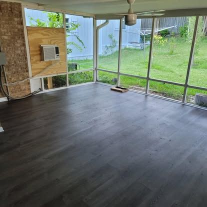 An indoor sunroom with dark wood floors, a brick wall, and a plywood panel housing a small air conditioning unit.
