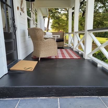A front porch with black painted flooring, a wicker chair, a red and white checkered rug, and a beige doormat.