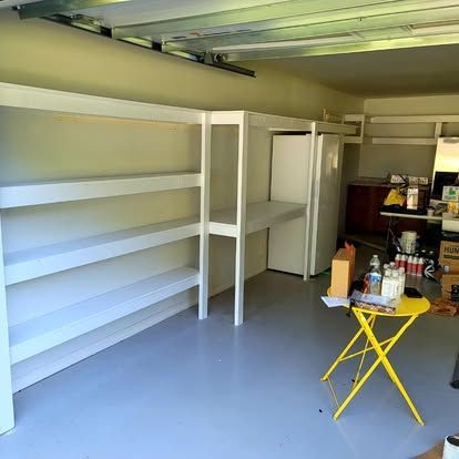 A garage with white wooden shelving units, a white refrigerator, and a small yellow folding table on a gray painted floor.