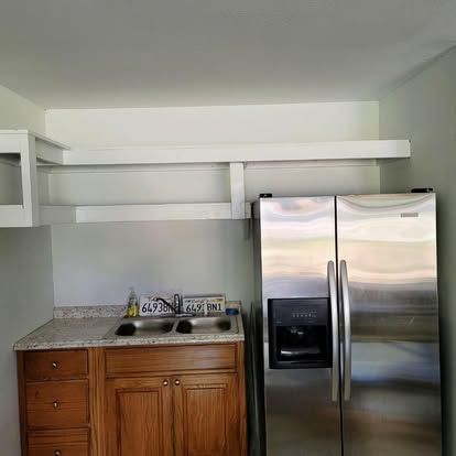 A stainless steel refrigerator next to a small wooden cabinet base with a sink and white wall-mounted shelves above.