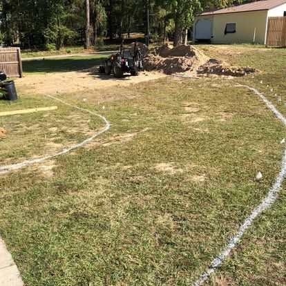 A grassy yard showing a white-sprayed curved outline for landscaping, a pile of dirt, and a small tractor in the background.