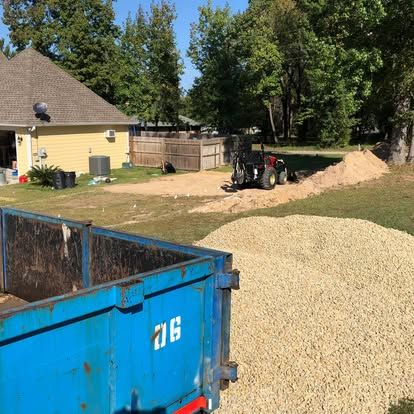 A blue dumpster in the foreground, with a tractor and piles of gravel in a residential yard near a house.