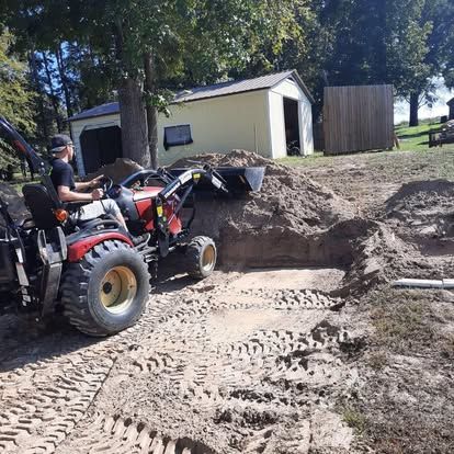 A person driving a red compact tractor with a front-end loader on a dirt lot next to a light-colored storage building.