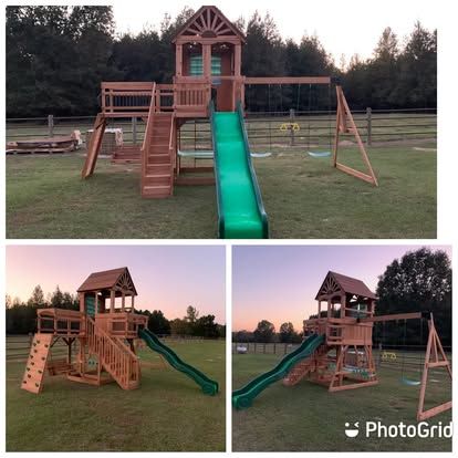 A wooden playground set with a green slide, climbing wall, swings, and a tower, situated on a grassy lawn at sunset.