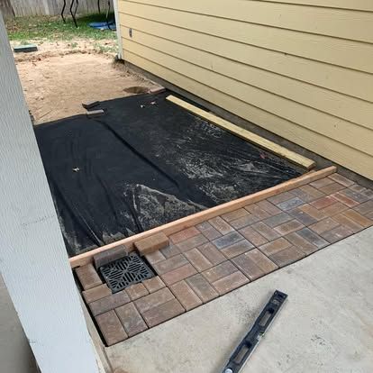 In-progress patio installation with pavers, a drainage grate, landscaping fabric, and wooden forms next to a yellow house.